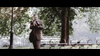 A man in a suit stands in a park with trees, benches, and a lamp post, holding a camera and looking through the viewfinder—reminiscent of scenes from The Caller (2008) DVD and other rare films.