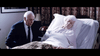 An elderly man in a suit sits by an elderly woman lying in bed with her eyes closed, covered by a quilt. The dim bedroom, flowers, and patterned curtains evoke the mood of "The Caller (2008) DVD.