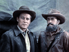 Two men in old-fashioned western attire stand side by side, one clean-shaven in a coat and the other bearded in a worn jacket, against a canvas backdrop. Scene from Kentucky Rifle (1955) DVD.