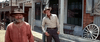 Two men in cowboy hats outside old western buildings evoke the American West; one in a red shirt stands ahead, another leans by a wagon wheel—relive classic western cinema with "These Thousand Hills (1959)" on DVD.