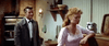 A man in a suit stands behind a woman in a light pink dress leaning on a kitchen counter. The rustic setting evokes scenes from classic films like These Thousand Hills (1959) DVD, with shelves and dishes enhancing the timeless atmosphere.