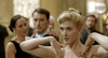 A blonde woman in a gold dress strikes a dramatic pose at a formal event, evoking scenes from the biographical drama "Nelly (2016)" DVD, while two people in black watch her with interest in an elegant, festive setting.