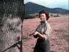 A woman in plaid and jeans stands by a tree outdoors, pistol in hand and alert—evoking the frontier spirit of The Lone Gun (1954) DVD, set against mountains and fields beneath a partly cloudy sky.