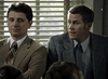 Two men in suits sit side by side indoors, sunlight streaming through blinds. One listens as the other discusses "The Investigation (2002) DVD," a gripping serial killer movie.