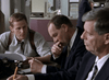 Three men in suits sit at a desk, deep in discussion, reviewing notes about The Investigation (2002) DVD. Papers and a pencil are on the desk, with office equipment visible in the background.