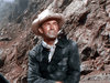 A man in a light cowboy hat, black jacket, and white neckerchief sits outdoors against mountains in the sunlight, evoking classic westerns like The Cariboo Trail (1950) DVD.