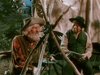 Two older men in vintage Western attire sit outdoors by a wooden structure, reminiscent of scenes from "The Cariboo Trail (1950) DVD," with a canvas tent and trees in the background evoking classic Technicolor westerns.