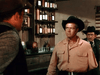 Three men in a western saloon—one in a pink shirt and black hat faces off with another in a vest, while a third man in a suit looks on. Bottles and glasses behind them recall classic scenes from The Cariboo Trail (1950) DVD.