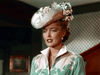 A woman in a vintage light green floral dress and a flowered hat stands indoors, evoking the timeless glamour captured in "The Cariboo Trail (1950) DVD," amid patterned wallpaper.