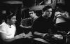 Four people are indoors in a black-and-white scene; three serious men sit around a table as a woman stands, partly smiling. The mood resembles a classic POW escape drama like "Break to Freedom (1953)" on DVD.