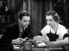 A man and a woman sit at a table, with the woman reading a paper as the man watches—a classic black-and-white scene from The Fighting Westerner (1935) DVD starring Randolph Scott.