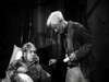 An older man with white hair holds a rolled-up paper beside a bed, as another man looks on with concern—this black and white scene captures the classic feel of vintage westerns like The Fighting Westerner (1935) DVD.