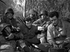 Five soldiers in uniform sit together in a jungle—one holding money, others relaxing or eating—in an intense scene from "Suicide Battalion (1958)," a classic World War II action film now available on DVD.