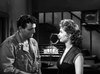 A man in uniform and a woman in a dark dress stand seriously in a dim room with a staircase and lamp, evoking the intense mood found in classic films like Suicide Battalion (1958) DVD.
