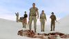Five people in desert gear stand on white sand, facing a tattered cloth—a scene from The Bone Snatcher (2003) DVD, a rare early 2000s horror film with South African creature feature vibes. A vehicle with a mounted gun looms nearby under the clear sky.