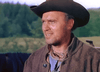 A man in a dark cowboy hat and brown jacket stands outdoors with a slight smile, evoking a scene from the Powder River (1953) DVD, against a backdrop of blurred greenery and trees.