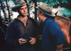 Two men in cowboy hats talk near a horse outdoors. One holds an object as the other in a blue shirt leans in. With trees and sky behind, it echoes a scene from the classic western "Powder River (1953)" DVD.