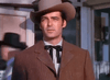 A man in a brown suit, black bowtie, and cowboy hat stands outside with a serious look—a scene straight from the Powder River (1953) DVD.