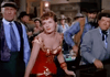 A woman in a red, off-the-shoulder dress stands at a saloon bar, surrounded by men in old Western attire and lively rustic decor—just like a scene from the classic Powder River (1953) DVD.