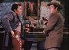 Two men in Western attire stand at a bar, one adjusting his holster, the other holding a drink. Bottles and a painting fill the backdrop—a scene straight from Powder River (1953) DVD, ideal for fans of classic westerns.
