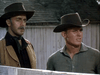 Two men in cowboy hats stand by a wooden fence, channeling the classic western vibe of "Gun Belt (1953) DVD." One wears a brown jacket and scarf, the other a plain shirt, with a building seen in the background.