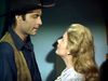 A man in a cowboy hat and vest stands facing a woman with long blonde hair, both locked in an intense gaze under soft lighting—evoking the timeless allure of classic scenes found in the “Gun Belt (1953) DVD.”.
