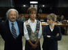 Three people stand in a courtroom: a suited man on the left, a woman in a patterned vest at center, and another with crossed arms on the right—a scene reminiscent of Judgement Day: The Ellie Nesler Story (1999) DVD.