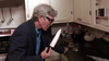 An older man in glasses and a dark jacket stands in the kitchen, holding a large knife near a counter with a drink—a scene straight from "Just What the Doctor Ordered (2021) DVD.