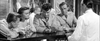 Four people in military uniforms and one man in white sit at a bar, appearing deep in thought or conversation with drinks on the counter. Blurred patrons and decor fill the background. This black-and-white image evokes the atmosphere found in rare hard to find movies, reminiscent of classic WWII drama DVDs such as “Bitter Victory (1957) DVD.”.