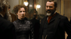 A woman in a high-necked, dark plaid Victorian dress converses with a bearded man in a suit with a red cravat at a formal gathering—evoking scenes from the "Madame Nobel (2014) DVD.