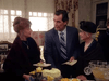 Three people—two older women in formal wear and a man in a suit holding an envelope—stand together at a gathering, evoking family drama themes seen in the Promise (1986) DVD. A table with cake, punch bowl, and fruit sits in front.