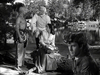 Four people in period attire gather by a lake and trees, reminiscent of a classic frontier scene. Featured on The Iroquois Trail (1950) DVD, characters include a uniformed man, woman with papers, shirtless man with beads, and one in a fringe jacket and coonskin cap.