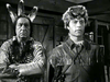 Two men in period costumes face each other indoors in this black-and-white scene from The Iroquois Trail (1950) DVD—one dressed in Native American attire with a feathered headdress, the other in a fringed jacket and coonskin cap.