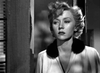 A woman stands in a doorway, short curled hair framing her contemplative face as she wears a robe with a dark fur collar—a single tear on her cheek. Experience the timeless drama of "In A Lonely Place (1950)" on DVD, captured in striking black and white.