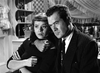 A man in a suit holds a concerned woman close in front of curtains and shelves. This black-and-white scene from "In A Lonely Place (1950) DVD" evokes classic Humphrey Bogart movies, ideal for fans of vintage cinema on DVD.
