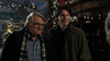 Two men stand indoors, smiling and chatting. Behind them are Christmas decorations—garlands and ornaments—that create a festive atmosphere reminiscent of a classic holiday film. In the background, shelves display various items, including DVDs such as “When Angels Come To Town (2004)” available on DVD.
