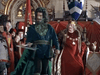 A man with green hair and beard, dressed in green, stands gripping an axe in a medieval hall as a crowned king in a red robe looks on, surrounded by onlookers—a vivid scene reminiscent of the Arthurian legend depicted in “Sir Gawain and the Green Knight (2021) DVD,” now available among rare classic movies on DVD.