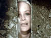 A woman with blonde hair and blue eyes gazes through a narrow gap between two stone walls, her face partially lit and her expression filled with hope or longing, evoking scenes from the film “Sir Gawain and the Green Knight (2021)” on DVD.
