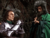 A knight in silver armor sits with hands clasped, looking somber, while a bearded man in a green cloak and fur headdress stands beside him holding a large axe—both set against a rocky backdrop, evocative of an iconic scene from Arthurian legend. This powerful imagery captures the timeless allure found in rare classic movies on DVD such as *Sir Gawain and the Green Knight (2021) DVD*.