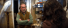 An older man with crossed arms stands in a cluttered shop across from a woman with curly hair, surrounded by shelves of boxes, rare movies on DVD—including “Tuesday (2023)”—and taxidermy animals.