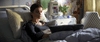 A woman with short hair lies in a hospital bed with an oxygen tube in her nose, gazing ahead thoughtfully, reminiscent of a scene from the “Tuesday (2023) DVD.” Medical equipment and a patterned chair can be seen in the background.