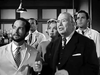 Four people, three in lab coats and one in a suit, stand closely together in a laboratory, their serious, concerned expressions reflecting the mood of a tense scene. Laboratory equipment and shelves fill the background, evoking the distinctive atmosphere found in classic films like Monkey Business (1952) DVD—those rare, hard-to-find movies that authentically capture moments of suspense.