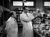Two men in lab coats stand in a laboratory. One man observes as the other carefully examines a flask filled with liquid. Lab equipment and glassware fill the workspace around them, capturing the zany spirit of “Monkey Business (1952)” on DVD, where madcap experiments and quirky characters are always just a scene away.