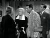 Four adults in formal 1950s attire stand indoors, engaged in a serious conversation. One woman faces away while the others look at her. This black and white scene evokes the intrigue and elegance found in classic films like “Monkey Business (1952)” on DVD—perfect for fans of rare, hard-to-find movies that capture a bygone era.