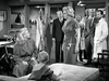 A group of adults, including men in suits and lab coats, stand in a room. Two women and a child are in the foreground, with one woman standing and appearing to speak intensely to the other, who is seated—perfectly capturing the vivid drama from classic films like Monkey Business (1952) on DVD.
