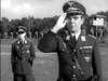 Two men in World War II-era German military uniforms stand outdoors; one is saluting in the foreground, while the other stands behind him. In the background, a watchtower and fence evoke scenes from “The Wooden Horse (1950)” DVD.
