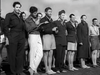 A group of men in military-style clothing and varied attire, including striped pants and shorts, stand together outdoors, posing for a photo on a sunny day—evoking a scene from “The Wooden Horse (1950)” DVD.