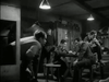 Six men sit and stand around a wooden table in a dimly lit room, engrossed in conversation and card games—a scene that echoes the rustic atmosphere depicted in “The Wooden Horse (1950) DVD,” where visible lockers and a hanging lamp overhead contribute to the film’s authentic, immersive setting.