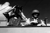 Two soldiers in helmets and vests stand aboard a ship; one peers through binoculars while the other gazes forward. Cables and smoke fill the background, creating a tense atmosphere reminiscent of a WWII underwater demolition thriller. Captured in black and white, the scene evokes the timeless style of classic films like The Frogmen (1951) on DVD.