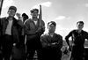 Five men in work clothes stand on a boat, gazing ahead beneath a bright, partly-cloudy sky. The serious and focused mood they convey as they face the camera—with water visible behind them—evokes classic WWII underwater demolition thrillers, perfectly capturing the timeless atmosphere found in rare, hard-to-find movies like *The Frogmen (1951)* DVD.